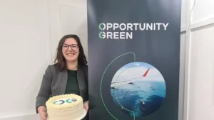 Photo of a woman wearing a grey jacket and glasses, smiling and holding a white circular cake that says OG on it. She stands next to a vertical banner taller than her with Opportunity Green written at the top and a circular image of a plane flying over ships in the sea.
