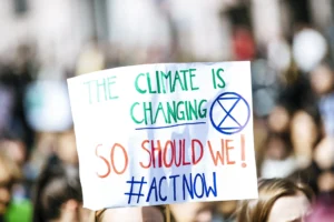 Banner held up at a protest, background is unfocused. The text reads: "The climate is changing, so should we! #ActNow". There is an hourglass shape in a circle, the logo of climate activism group Extinction Rebellion.