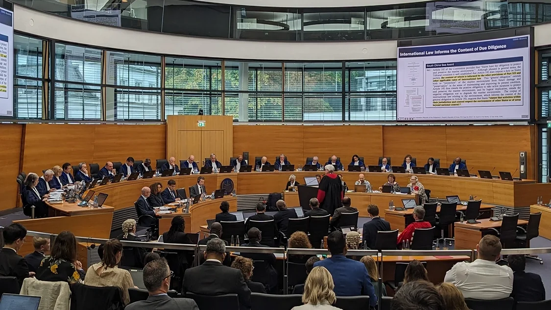 Image of ITLOS plenary, people in suits sitting in a circular room with wooden panelling, and a presentation projected onto a screen (not legible). Image of ITLOS plenary, people in suits sitting in a circular room with wooden panelling, and a presentation projected onto a screen (not legible).