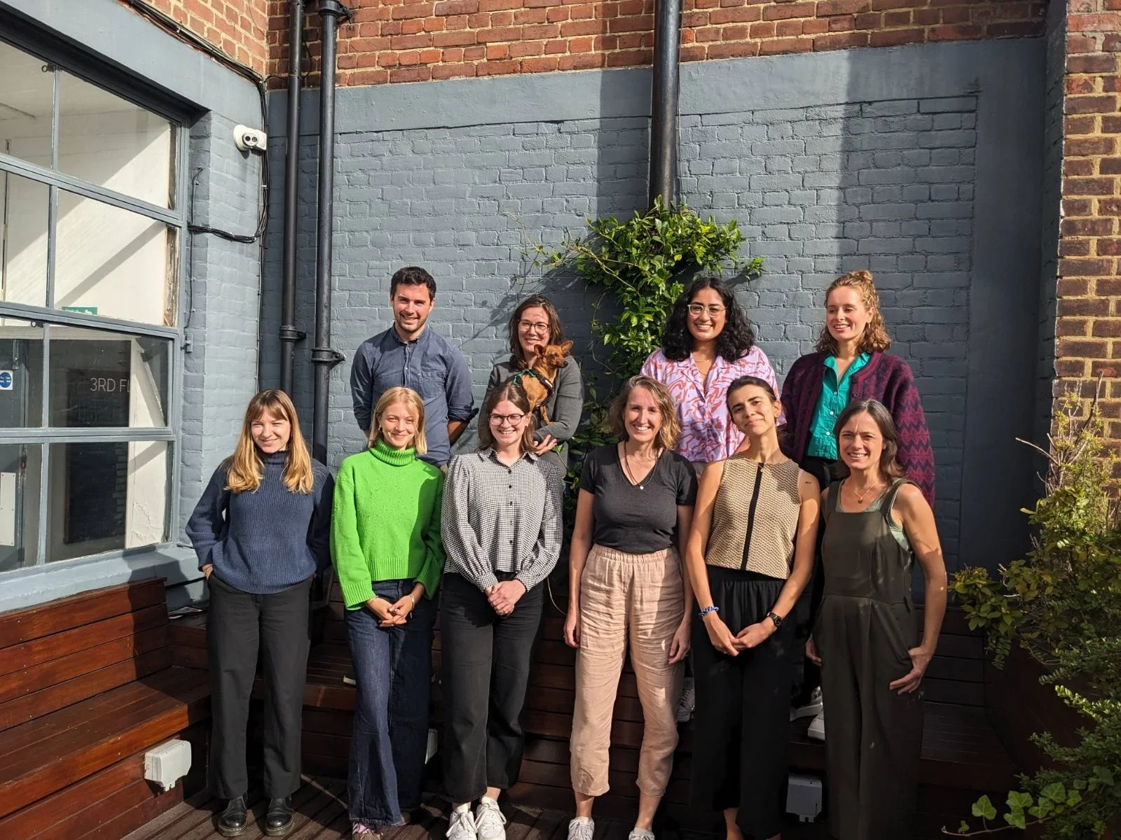 Photo of ten people in two rows posing for a team photo. It is sunny and they are on a balcony smiling. There is a brick wall painted grey and a plant behind them.