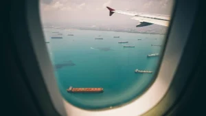 Photo taken through the window of an airplane. You see the sea below with ships in it and the left wing of the plane.