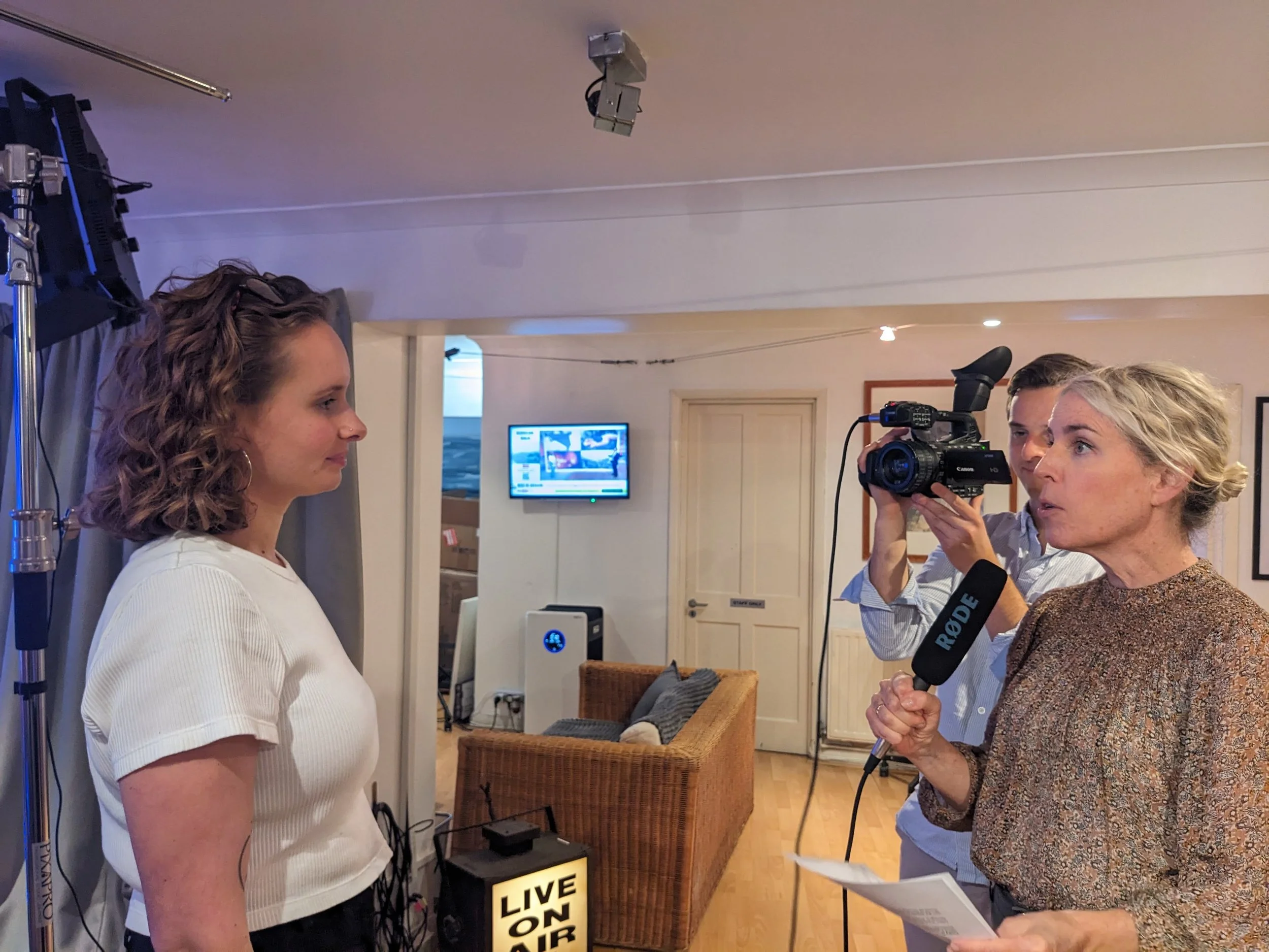 Two women facing each other inside. The one on the left is being interviewed by the one on the right. A man behind the woman on the right is holding a video camera towards the woman on the left.