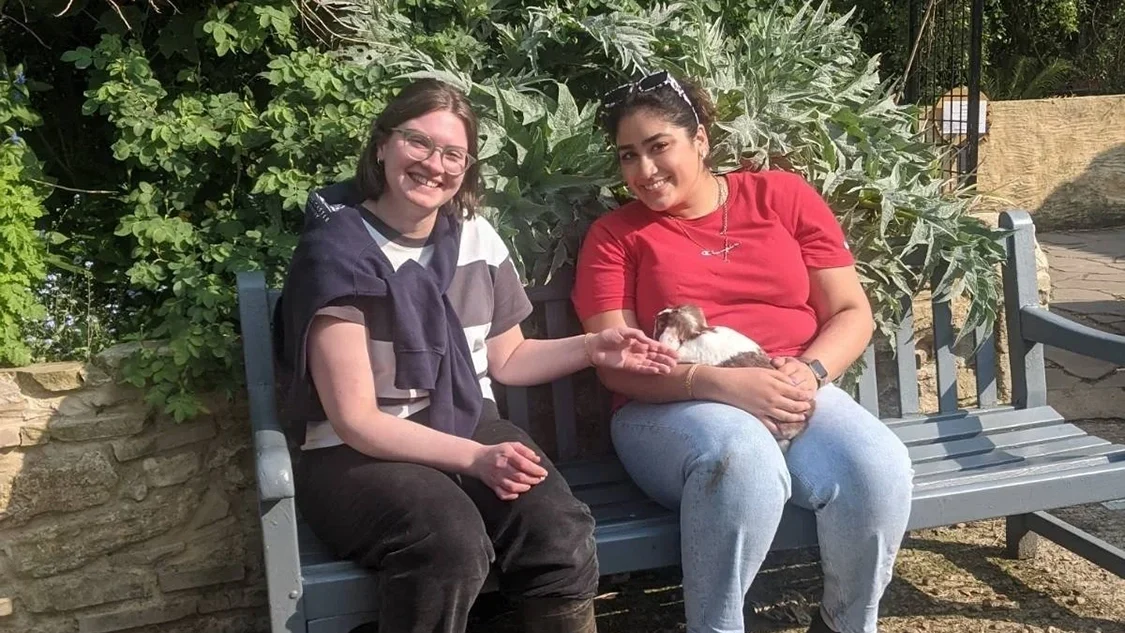 Two women sit on a bench, both smiling. The one on the left his a blue and white t-shirt on and a blue jumper tied around her neck, and black trousers. The woman on the right has a red t-shirt on and is holding a guinea pig. The one on the left is also stroking the guinea pig.