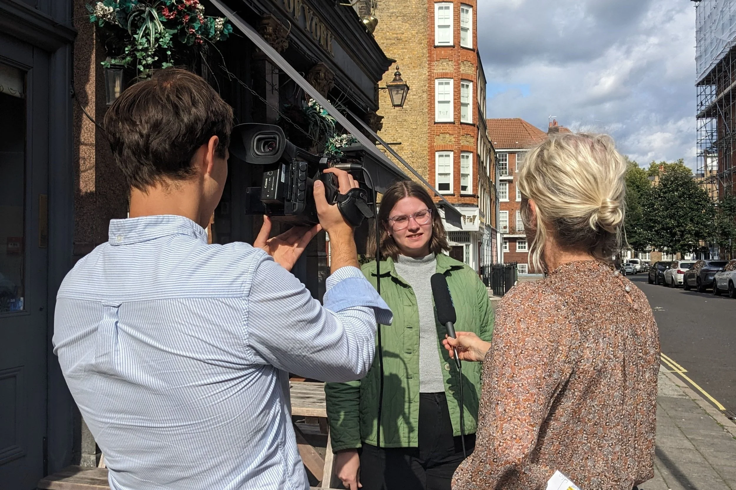 A woman in a green jacket looks forward and is speaking. A blond woman with her back turned holds a microphone towards the first woman, and a man with his back turned holds a camera towards her. It is an interview