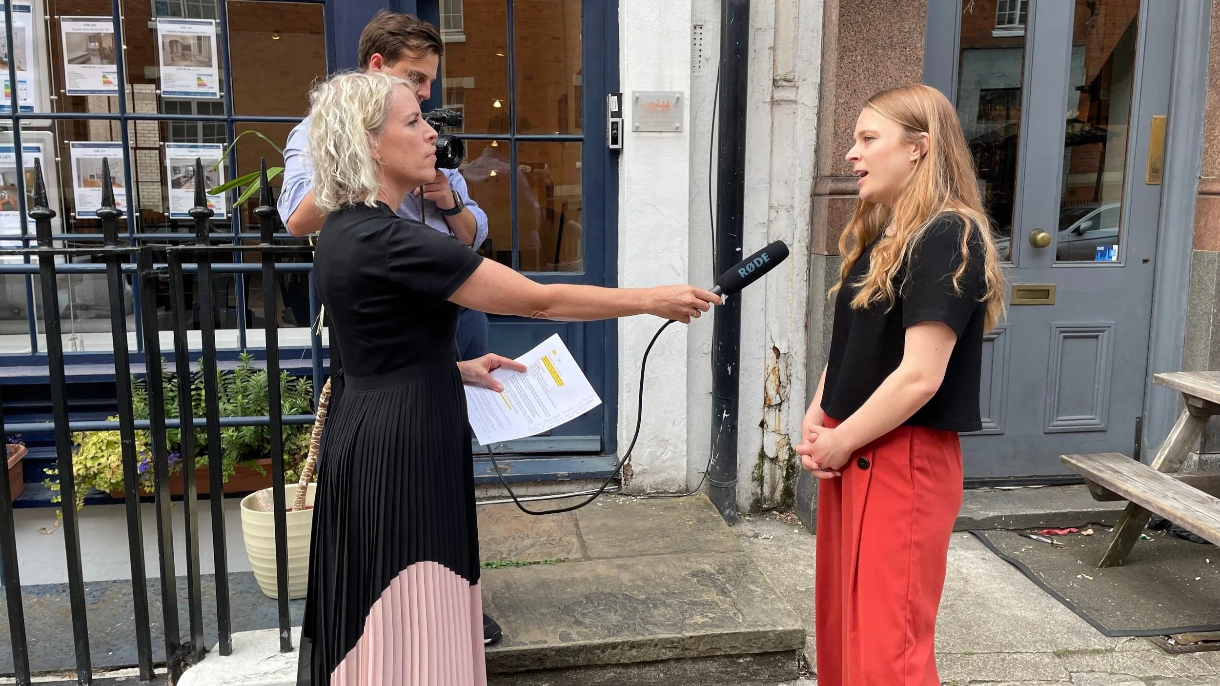 One women holds microphone and interviews another woman, standing on the street