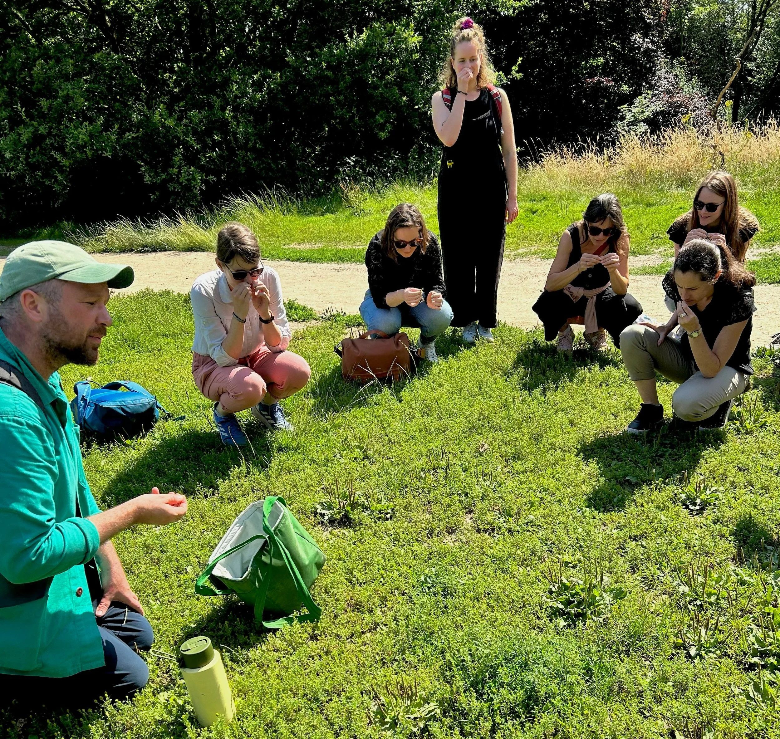 A man on the left in a green jacket and green hat is speaking, gesturing with his hand. Five women are crouching on the grass, smelling something in their hands. One woman is standing, smelling the same thing. The floor is grassy and it is sunny, there is a path in the background and trees.