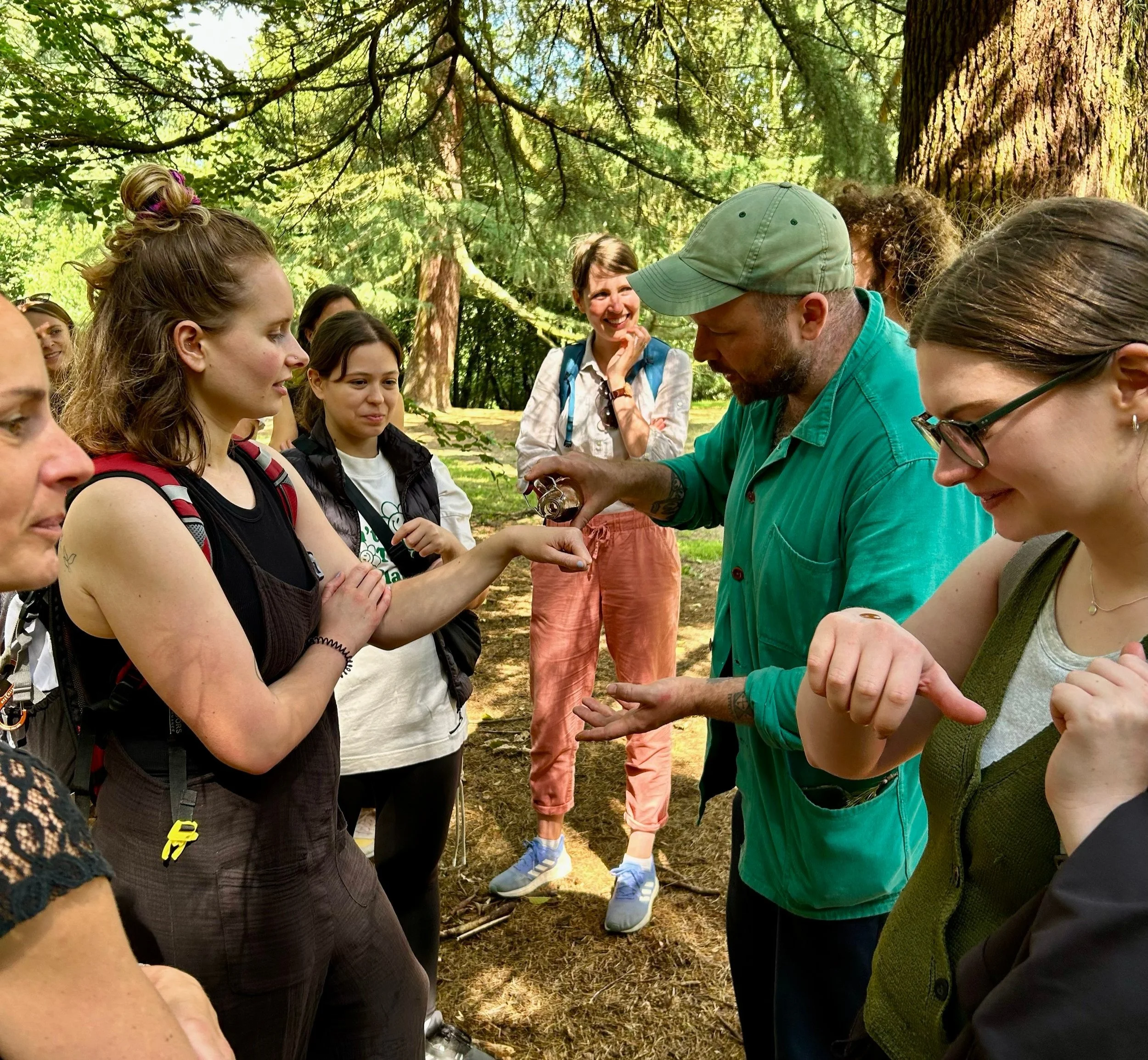 A man in a green hat and green jacket puts a drop of pine syrup on people's hands that they lick. There are trees and leaves in the background. The people are smiling and looking curious.