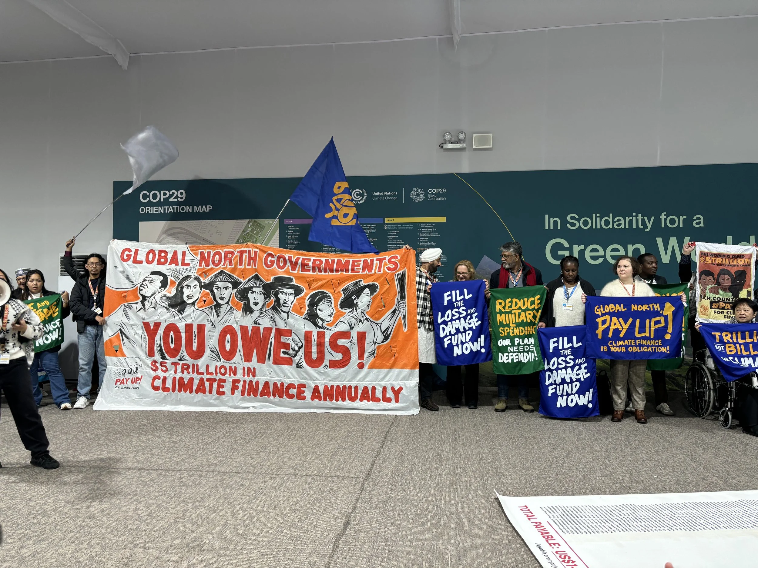 Activists at COP29 in Baku, Azerbaijan, with banners reading:
"Global north governments, you owe us! $5 trillion in climate finance annually."
"Fill the loss and damage fund now!"
"Reduce military spending, our planet needs defending"
"Global north pay up! Climate finance is your obligation!"