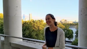 A woman with a black tshirt and grey cardigan smiles looking to the side. Behind her is a sunny London view of trees. There are two stone columns on either side of her.