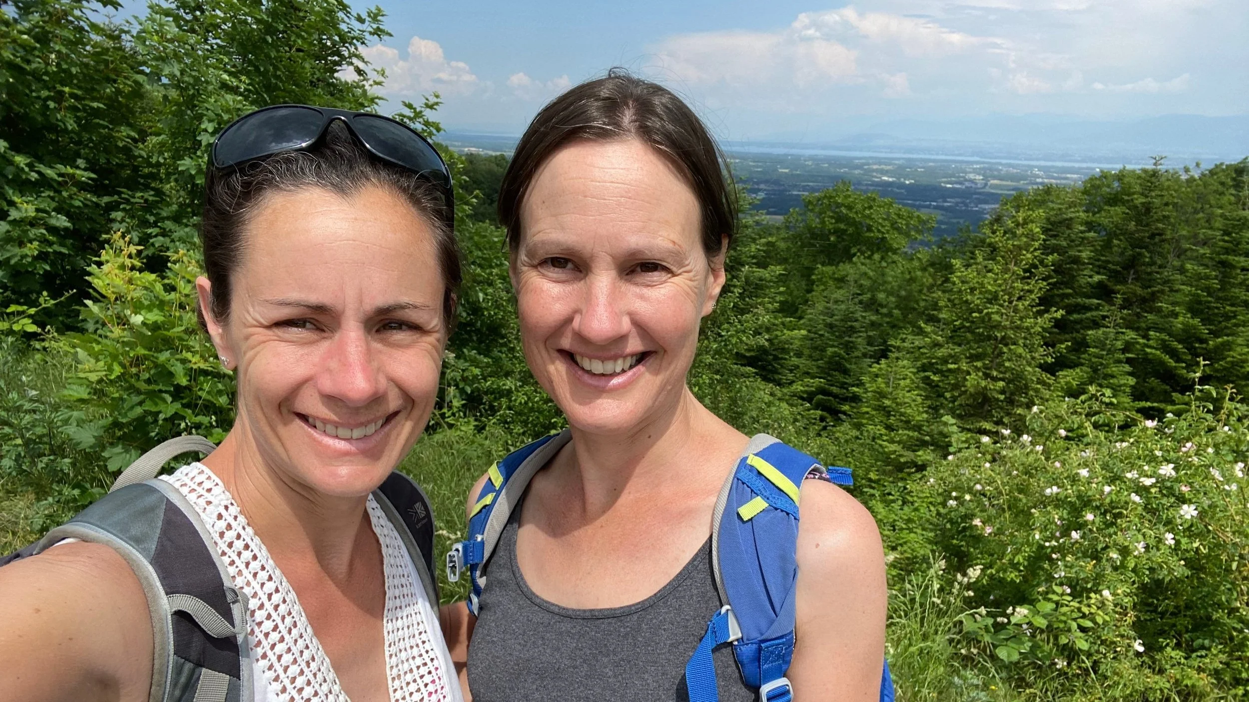 Two women standing close together with backpacks on and trees behind them smiling.