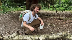 A man with curly hair in a white t-shirt and wearing a backpack crouches on a forest floor next to a fungus growing out of a log, and smiling.