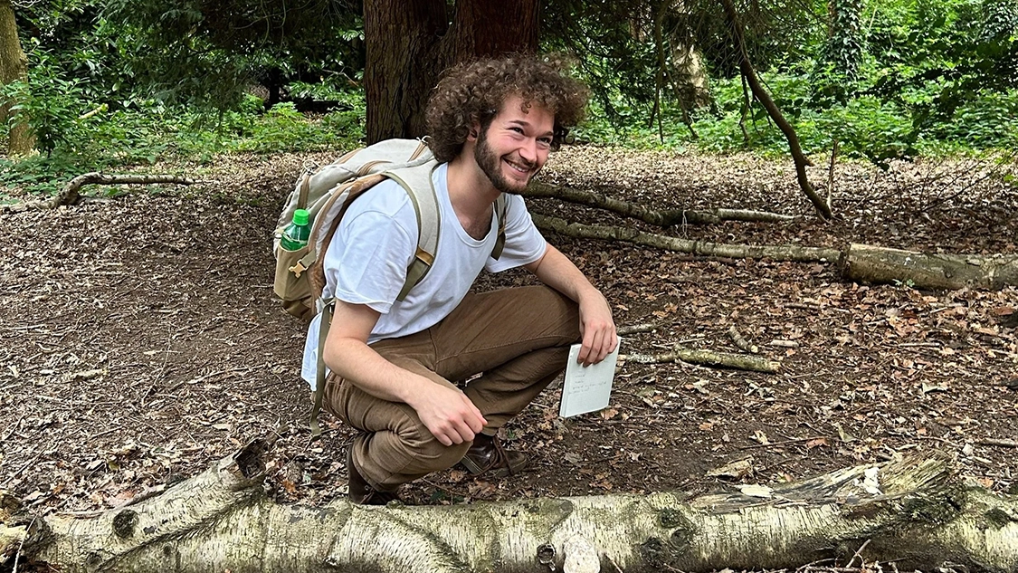 A man with curly hair in a white t-shirt and wearing a backpack crouches on a forest floor next to a fungus growing out of a log, and smiling. A man with curly hair in a white t-shirt and wearing a backpack crouches on a forest floor next to a fungus growing out of a log, and smiling.