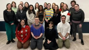 20 people pose for a photo smiling. They are in three rows. Some wear Christmas jumpers.