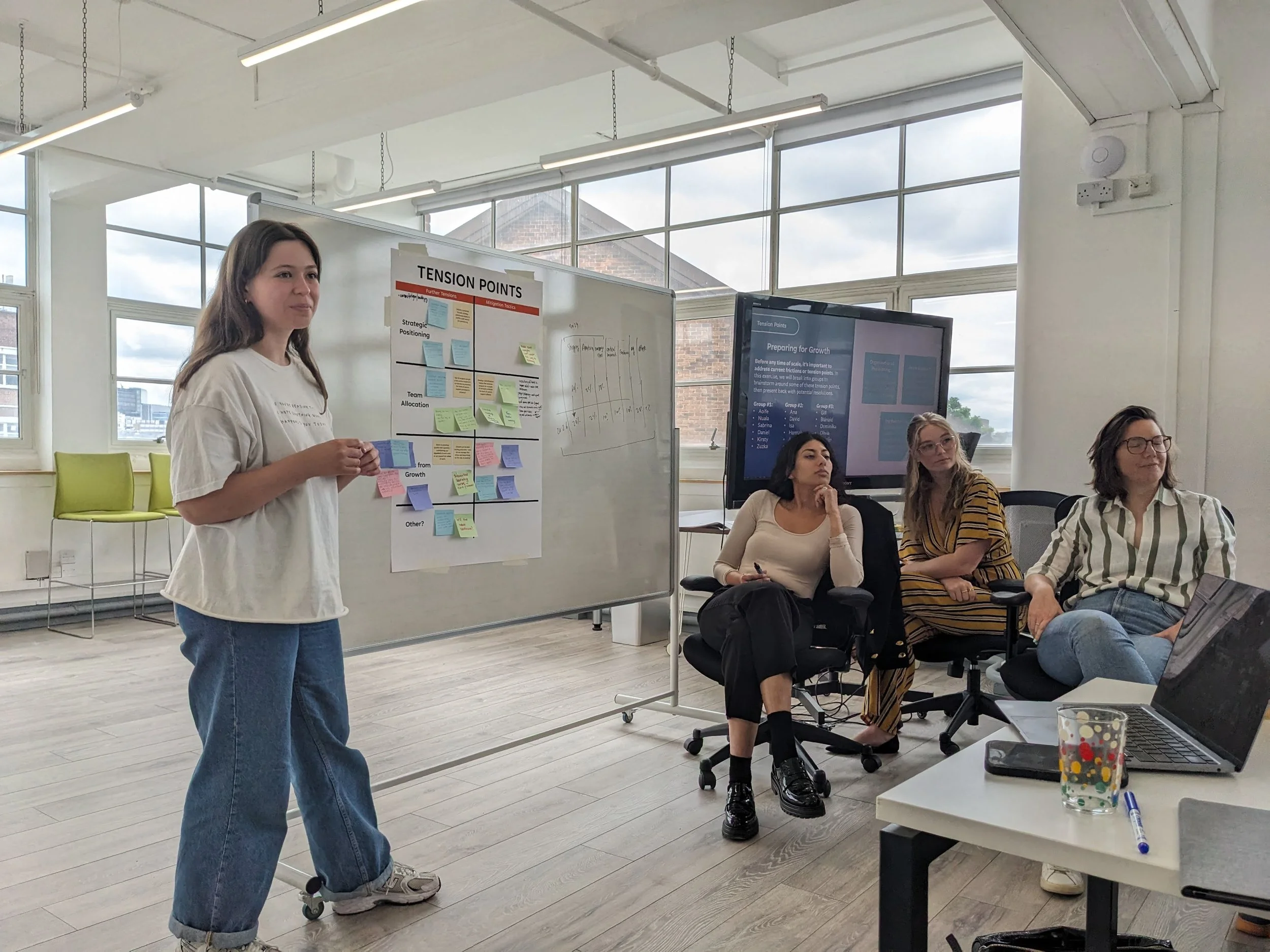 Photo of a woman on the left in jeans and a white shirt standing next to a board that has coloured post-it notes stuck to it and the heading "Tension points" at the top. She is giving a presentation. There are three other women sitting in chairs on the right in front of a television screen with a presentation on it.