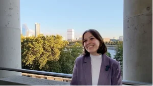 Picture of a woman in a purple jacket smiling with a sunny view of trees and the River Thames in London behind her, and two stone columns on either side.