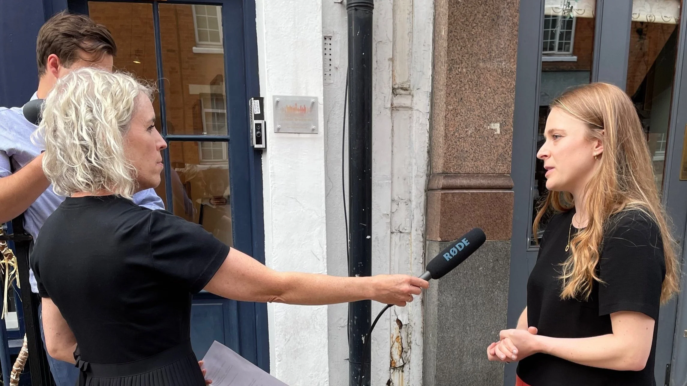 Two women both with black shirts. The one on the left holds a microphone out to the other who is speaking.