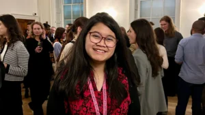 A woman with dark hair and glasses and a red patterned dress smiling. There are a lot of people behind her at a business party.