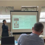 Woman in black leather jacket presenting from a projected slide, other staff members sit around tables watching.