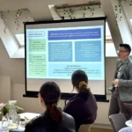 Person in grey jacket presenting from a projected slide, other staff members sit around tables watching.