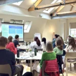 Woman in pink shirt presenting from a projected slide, other staff members sit around tables watching.