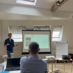 Man in blue shirt presenting from a projected slide, other staff members sit around tables watching.