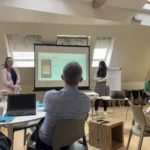 Two women, one with pink shirt one with white jumper and long dark hair, presenting from a projected slide, other staff members sit around tables watching.
