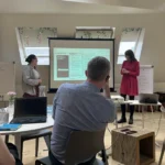 Two women, one in pink dress, one with grey shirt presenting from a projected slide, other staff members sit around tables watching.