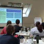 Woman in dark shirt presenting from a projected slide, other staff members sit around tables watching.