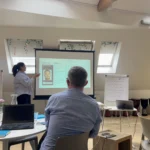 Woman in shirt presenting from a projected slide, other staff members sit around tables watching.