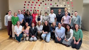 Photo of the Opportunity Green team, 23 staff members in total, some people kneeling, others standing. The floor is wooden and there is a spotted design on the wall behind them.