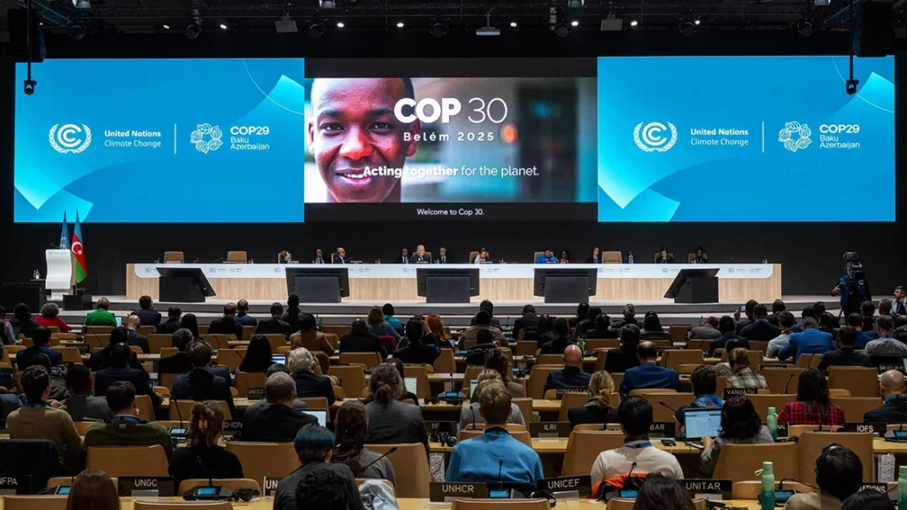 Image of people in a large room with screens at front. The two blue ones say United Nations Climate Change COP29 Baku Azerbaijan. The one in the middle has a picture of a man smiling on it and reads "COP30 Belém 2025, Acting together for the planet."