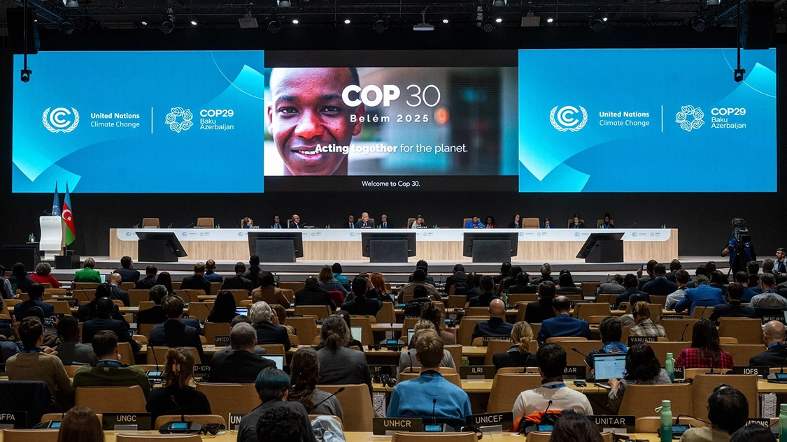 Image of people in a large room with screens at front. The two blue ones say United Nations Climate Change COP29 Baku Azerbaijan. The one in the middle has a picture of a man smiling on it and reads "COP30 Belém 2025, Acting together for the planet." Image of people in a large room with screens at front. The two blue ones say United Nations Climate Change COP29 Baku Azerbaijan. The one in the middle has a picture of a man smiling on it and reads "COP30 Belém 2025, Acting together for the planet."