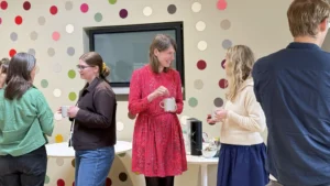 There are three women, the woman on the left wears a brown jumper adn her hair tied up and blue jeans. The woman in the middle is holding a mug and wearing a red dress with mid-length hair. She is speaking to the woman on the right who has blond hair, blue trousers and a white jumper. There are coloured circles on the wall in the background.