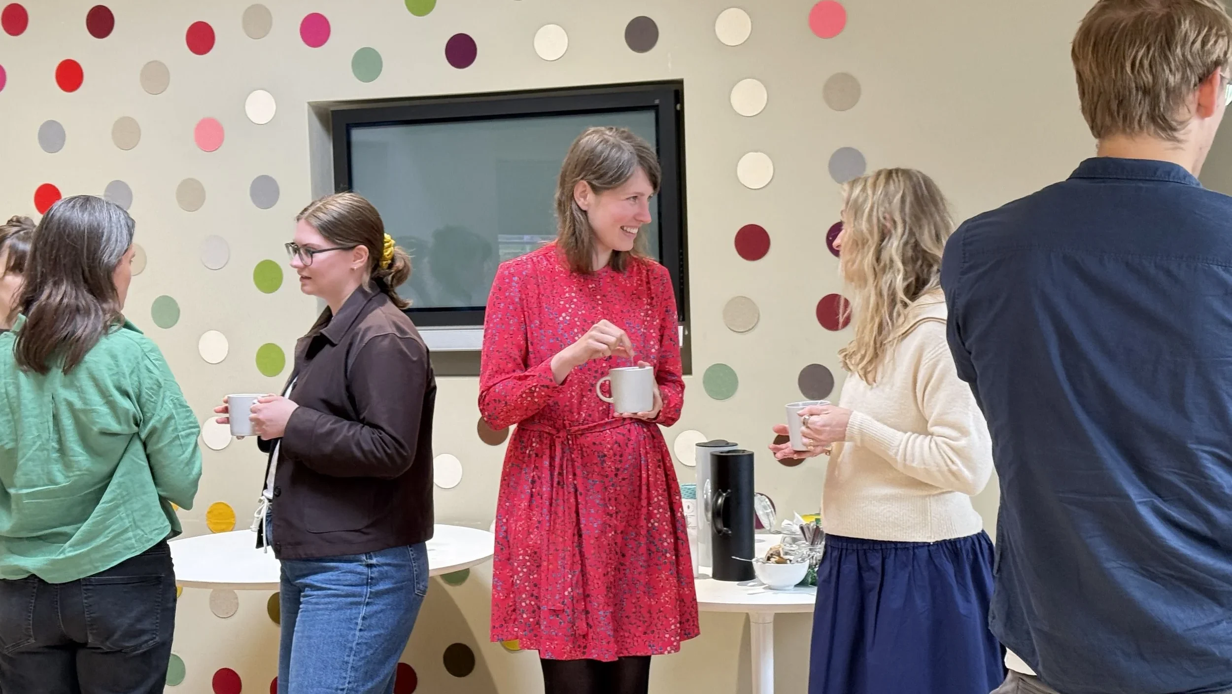 There are three women, the woman on the left wears a brown jumper adn her hair tied up and blue jeans. The woman in the middle is holding a mug and wearing a red dress with mid-length hair. She is speaking to the woman on the right who has blond hair, blue trousers and a white jumper. There are coloured circles on the wall in the background.