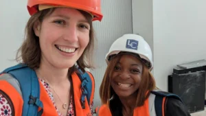 Photo of two women smiling in high visibility orange jackets and wearing hard hats.