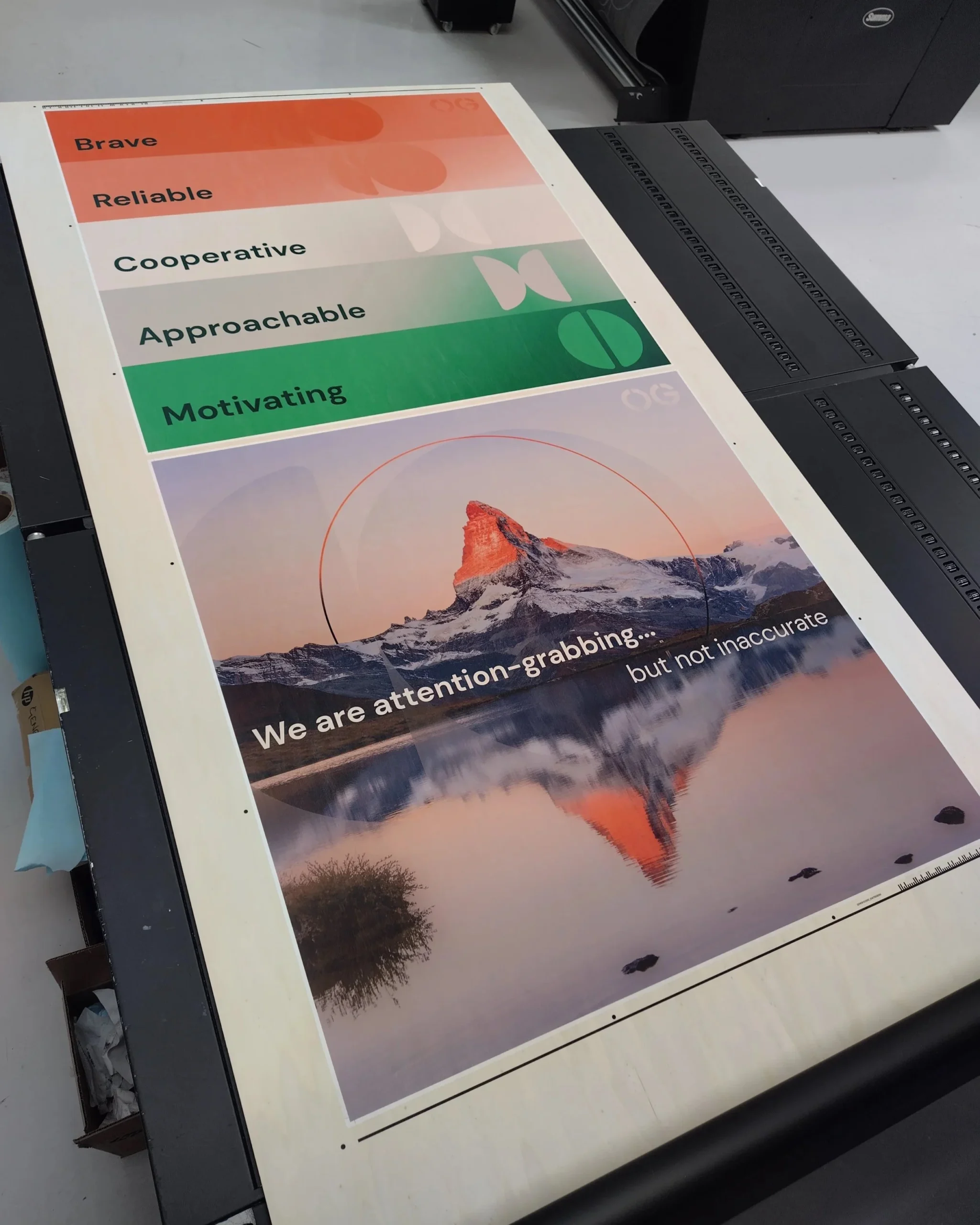 Image of two posters in a professional printers. One has an image of a mountain on it, and the slogan "We are attention grabbing... but not inaccurate". The other has five coloured lines (red, orange, cream, grey, green) with the words "Brave, Reliable, Cooperative, Approachable, Motivating" in each line. At the right of each line is a shape.