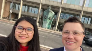 Two people outside the International Maritime Organization in London, which has a statue of a ship's front and a person on top of it in green/blue metal.