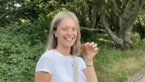 Woman in a white t-shirt with a forest behind her is smiling and holding up a red fungus in her left hand.