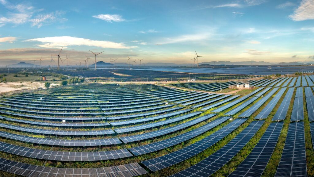 Many rows of solar panels in field with wind turbines and mountains in background