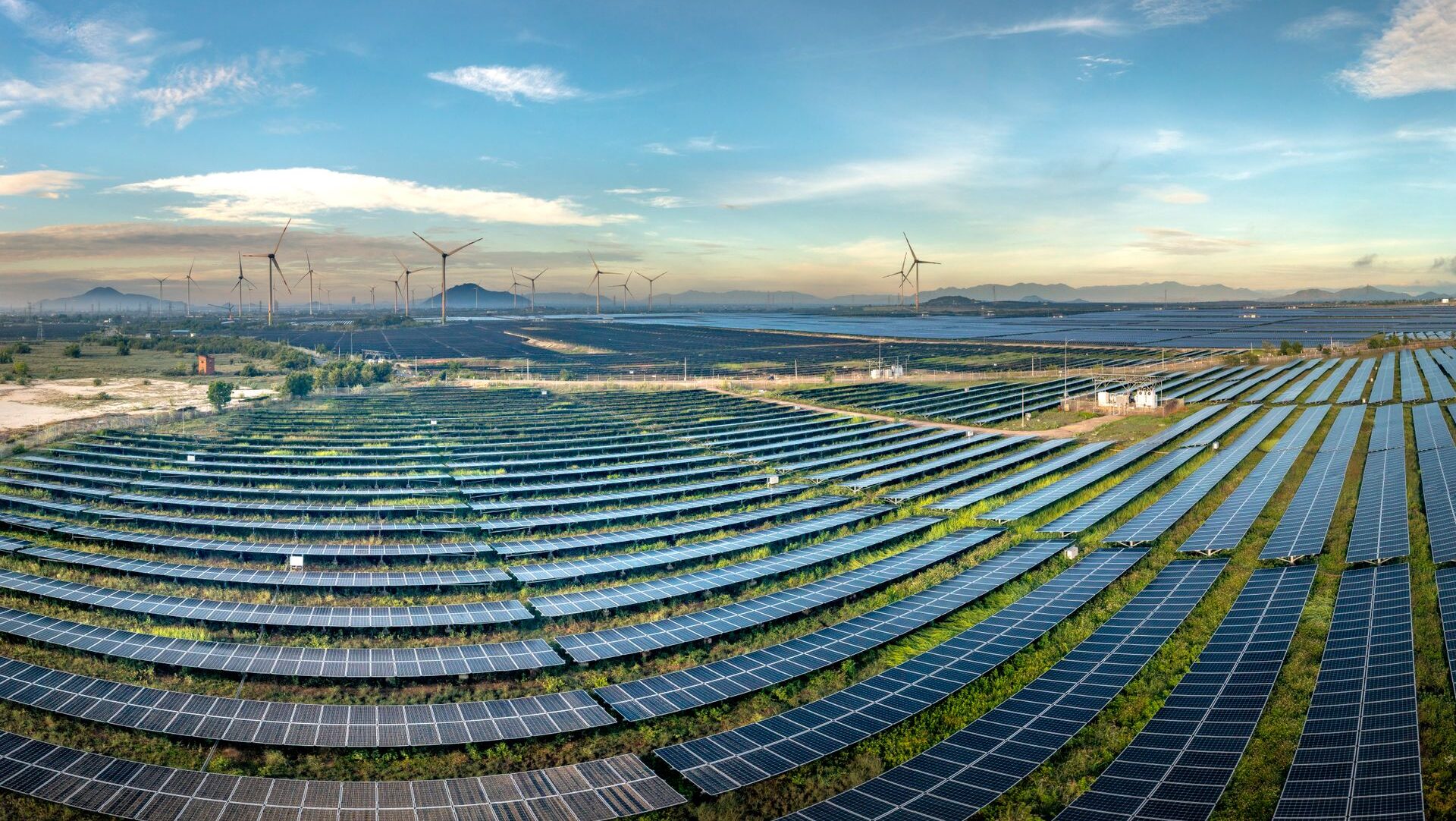 Many rows of solar panels in field with wind turbines and mountains in background
