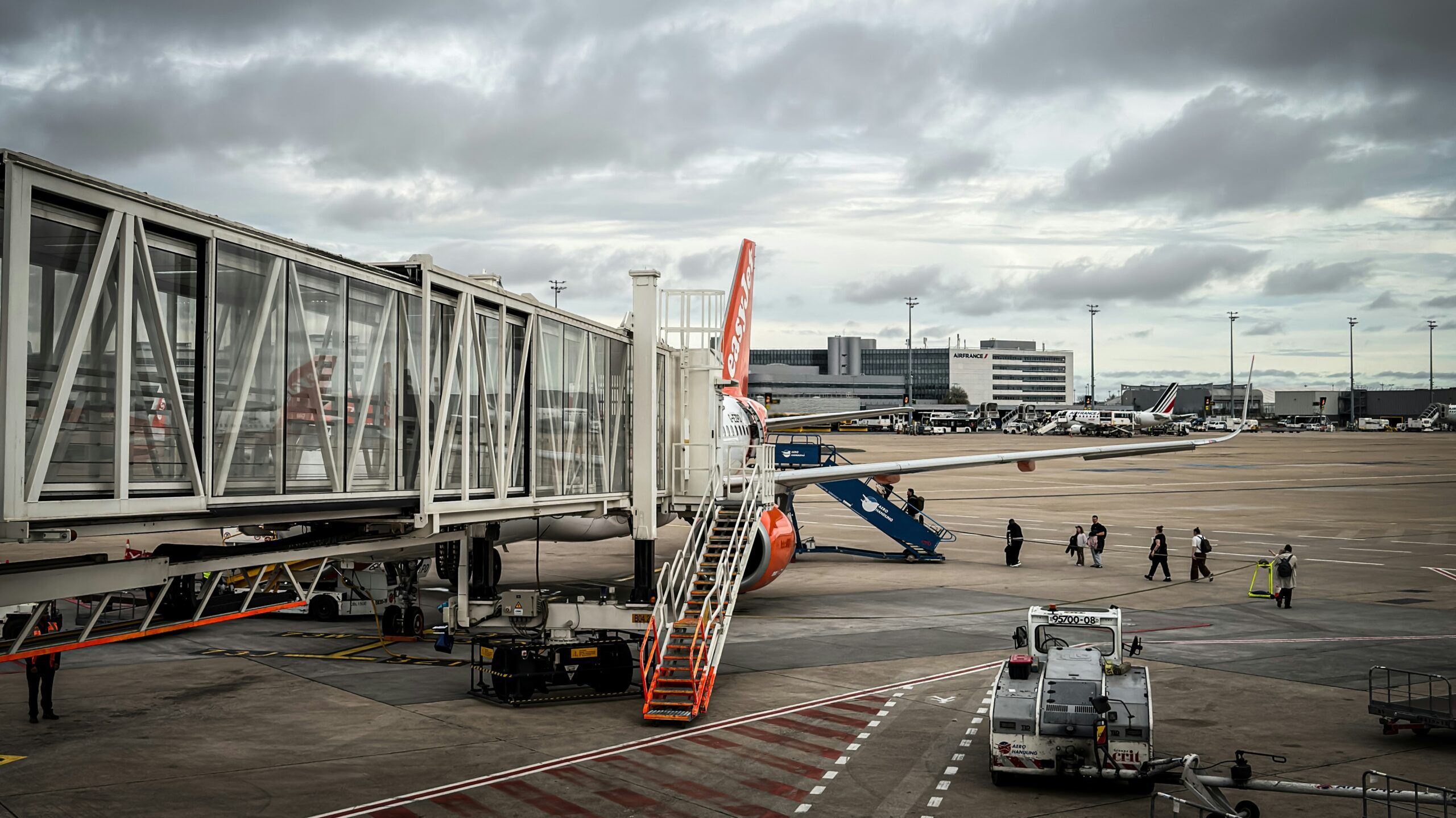 A plane parked at an airport gate attached to a glass jet bridge and boarding stairs on a cloudy tarmac with ground equipment and passengers walking below.