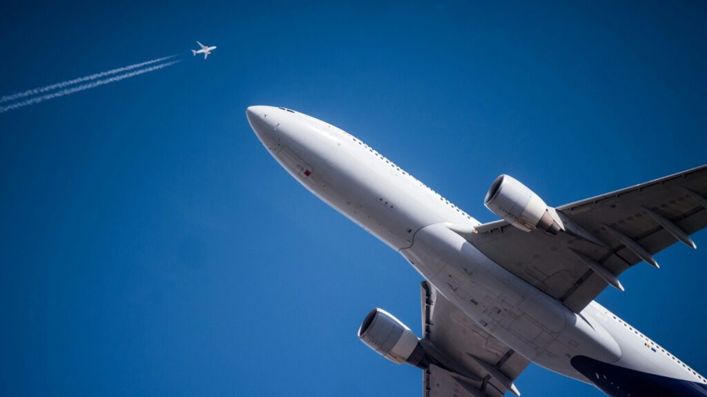Low-angle view of a white commercial airliner in flight against a bright blue sky, with a smaller airplane and contrail visible high in the background.