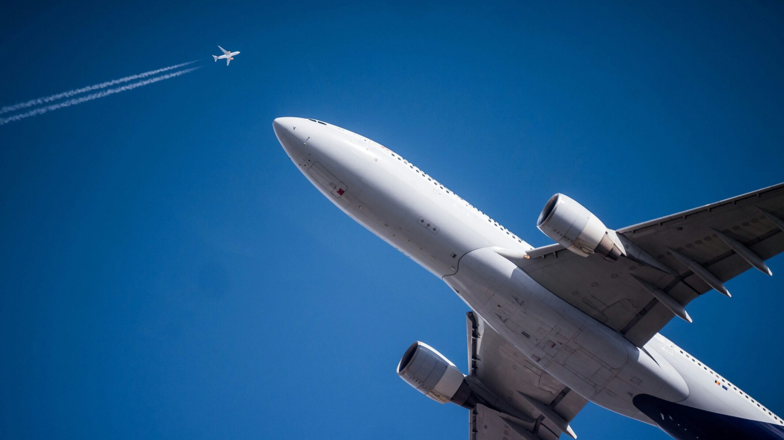 Low-angle view of a white commercial airliner in flight against a bright blue sky, with a smaller airplane and contrail visible high in the background.